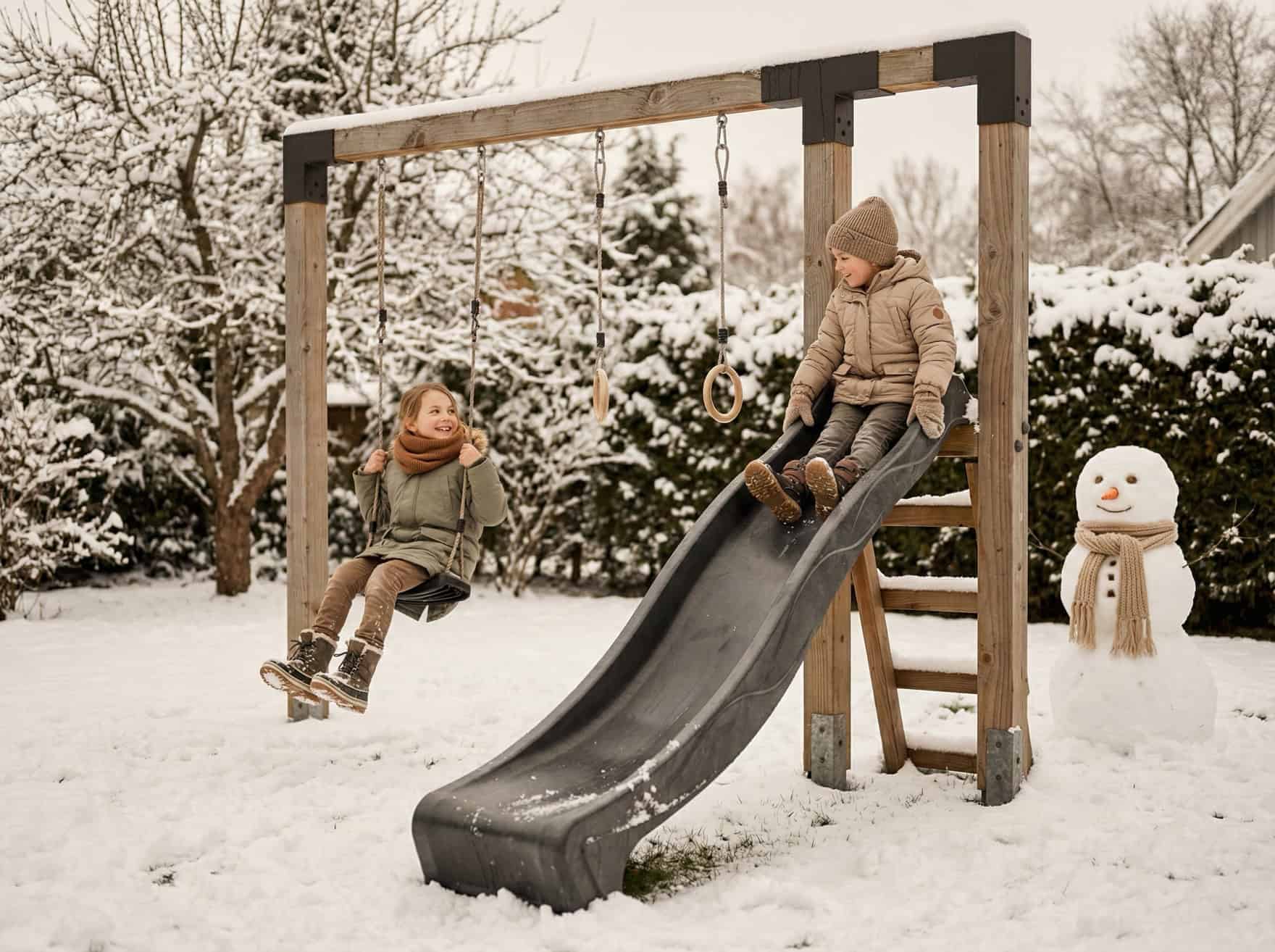 Twee kinderen spelen buiten op een houten speeltoestel met schommel, glijbaan en duikelrek van EXIT Toys in een besneeuwde tuin.